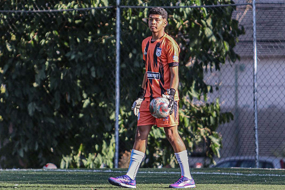 Goleiro Alisson Gabriel em campo durante treino, vestindo uniforme laranja e preto do Clube Vital SAF, segurando a bola com luvas e olhando à frente, com expressão concentrada, sobre gramado sintético e fundo de cerca e árvores.