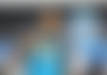 Fotografia colorida em plano médio, tirada em ângulo ligeiramente baixo, do zagueiro Wallace Santos com a camisa oficial do Grêmio (listrada em azul celeste, preto e branco). Ele está em pé em uma sala de imprensa ou área mista, olhando para a câmera com um sorriso largo, bigode e cavanhaque, e fazendo um sinal de 'joia' com o polegar direito levantado. O escudo do Grêmio e patrocinadores como 'OLINA' e 'Coral' estão visíveis na camisa da marca New Balance. O fundo é uma parede cinza escuro com o grande brasão do Grêmio e a data '1903' acima, além de banners desfocados de outros patrocinadores.