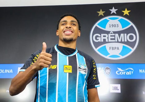 Fotografia colorida em plano médio, tirada em ângulo ligeiramente baixo, do zagueiro Wallace Santos com a camisa oficial do Grêmio (listrada em azul celeste, preto e branco). Ele está em pé em uma sala de imprensa ou área mista, olhando para a câmera com um sorriso largo, bigode e cavanhaque, e fazendo um sinal de 'joia' com o polegar direito levantado. O escudo do Grêmio e patrocinadores como 'OLINA' e 'Coral' estão visíveis na camisa da marca New Balance. O fundo é uma parede cinza escuro com o grande brasão do Grêmio e a data '1903' acima, além de banners desfocados de outros patrocinadores.
