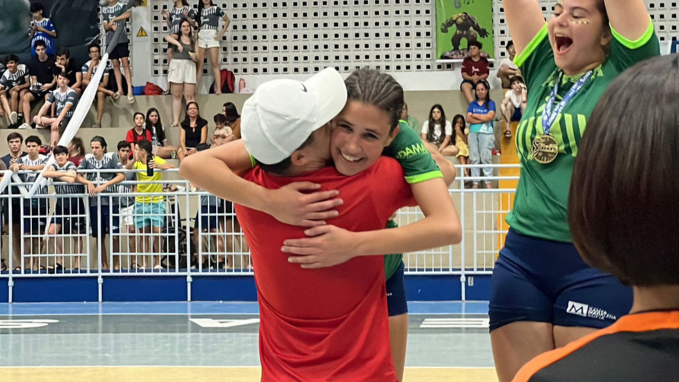 Bia Gomes, sorridente e emocionada, abraça seu pai com força após conquistar o título no futsal. Ela veste uniforme verde com detalhes em amarelo, enquanto uma colega de equipe comemora ao fundo com medalha no pescoço e braços erguidos. Ao redor, a arquibancada está cheia de torcedores e colegas que assistem à cena de celebração.
