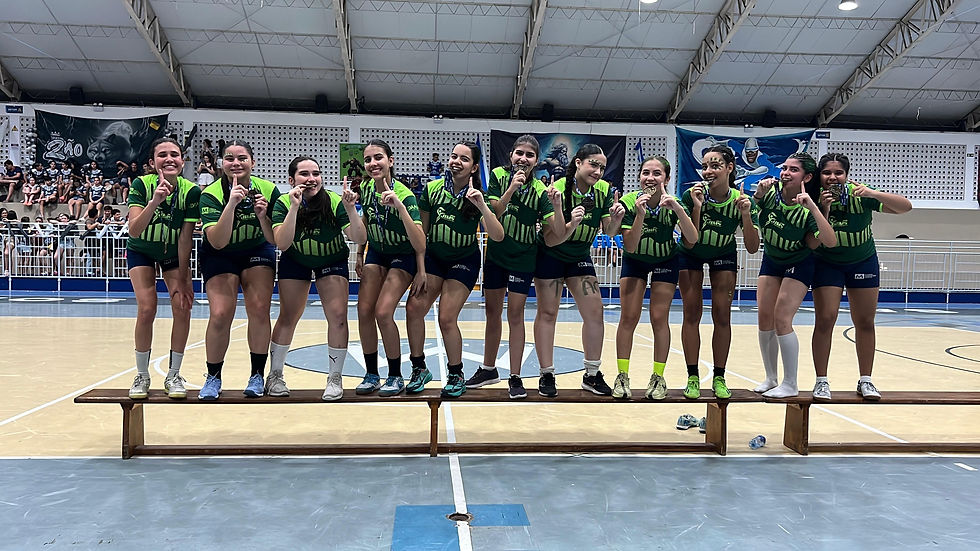 Bia Gomes posa sorridente ao lado de suas companheiras de equipe, todas vestindo o uniforme verde com detalhes em azul do Colégio Madalena Sofia. As atletas estão alinhadas sobre um banco no centro da quadra, comemorando o título escolar com medalhas na boca e fazendo o gesto de “número 1” com os dedos. Ao fundo, a arquibancada está cheia de colegas e torcedores, celebrando a vitória em uma competição escolar animada e muito bem organizada em Maceió.