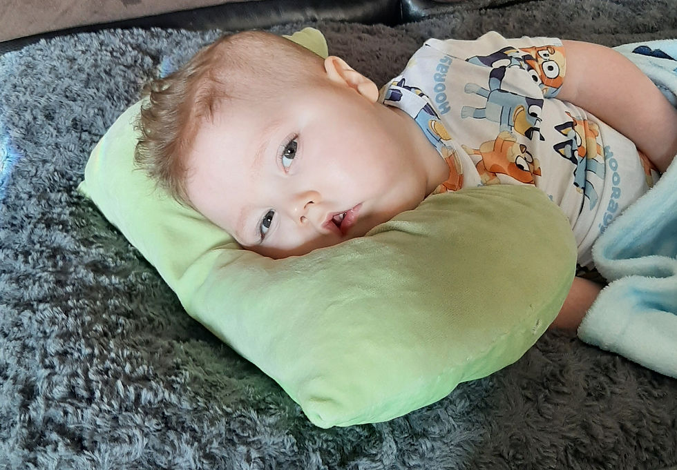 Baby lying on a green pillow, wearing a cartoon print onesie saying "Hooray," on a gray fluffy blanket. Calm and relaxed mood.