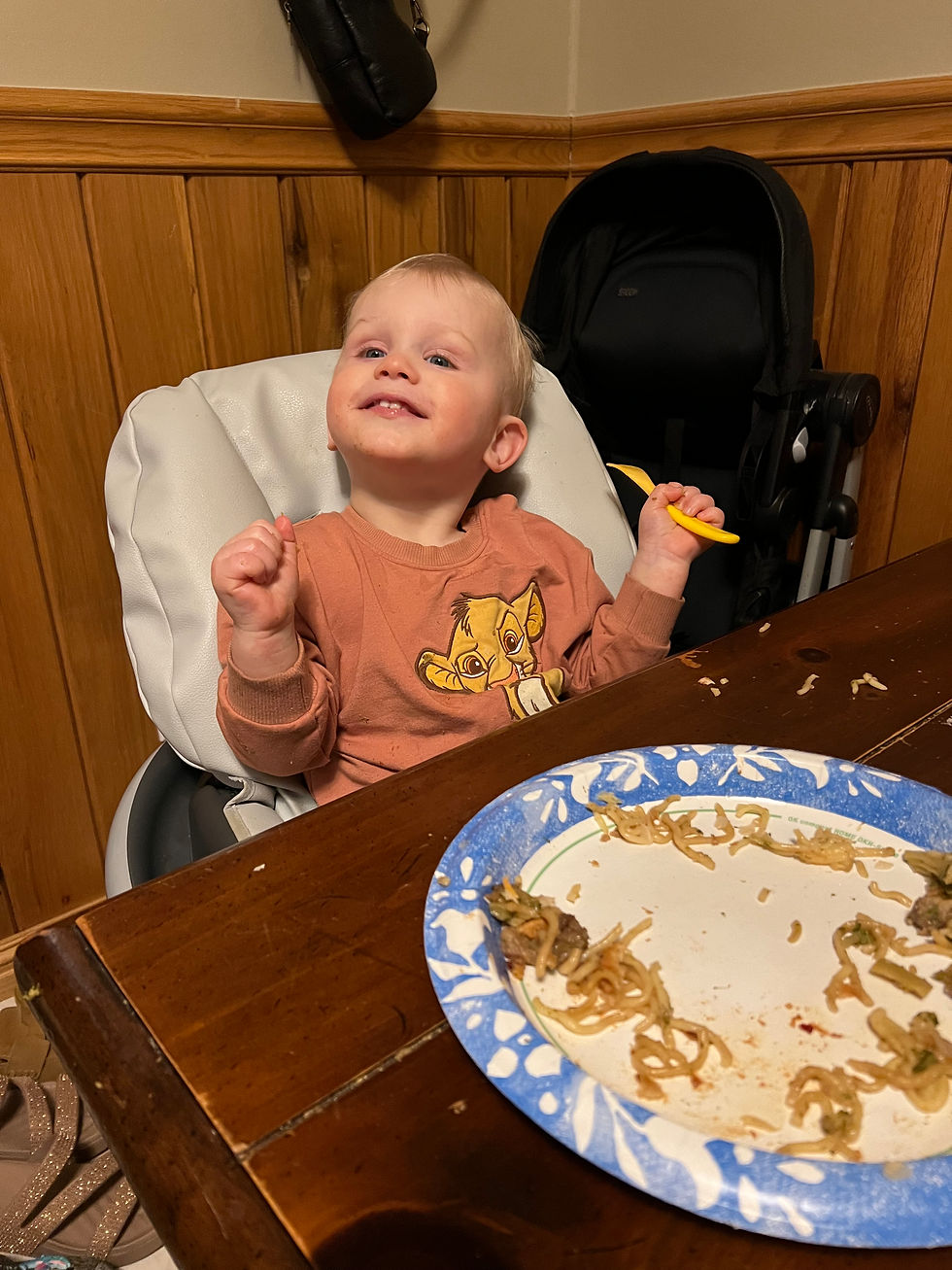 A smiling toddler in a high chair holds a yellow fork. A plate with noodles is on the wooden table. Wearing a brown shirt with a lion design.