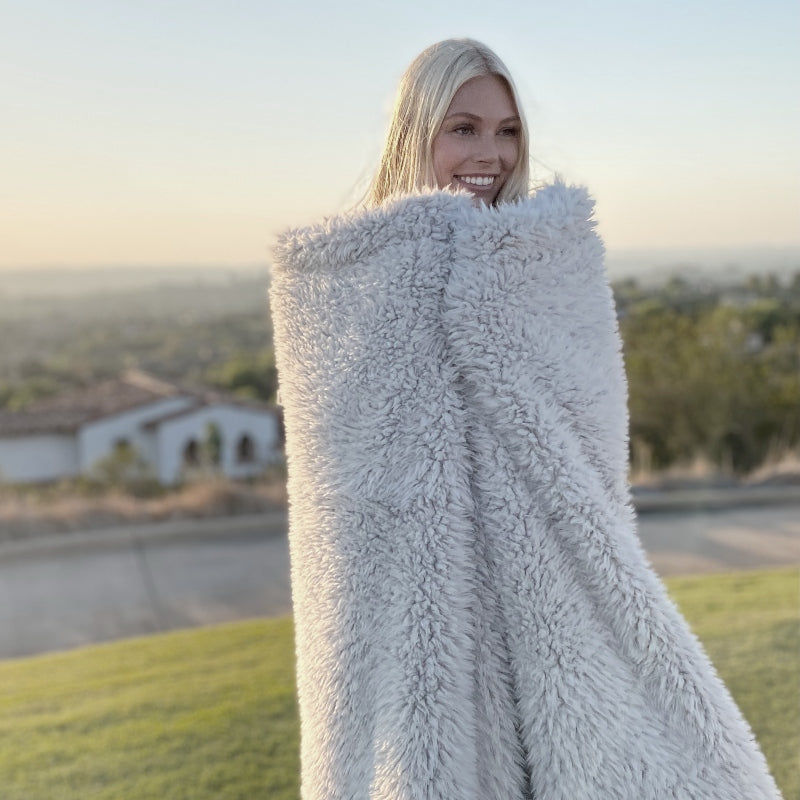 Blonde woman wrapped in a fluffy gray blanket, smiling outdoors, sunny landscape.