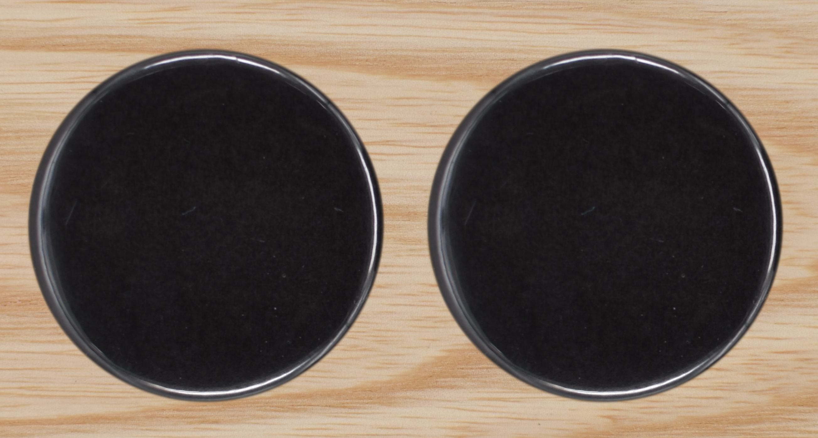 Two black, glossy circles sit on a light wooden textured background Khaam Fefalene