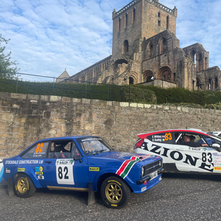 Rally cars are parked in the morning sunshine, with Jedburgh Abbey in the background
