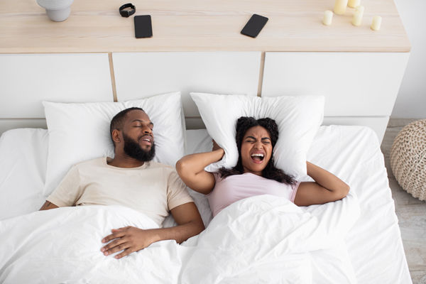 husband and wife in bed, man is snoring and woman is covering her ears from loud snoring