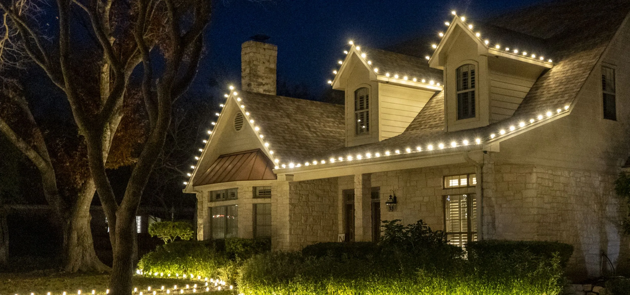 Two-story home with warm white roofline and dormer lights and lit landscaping