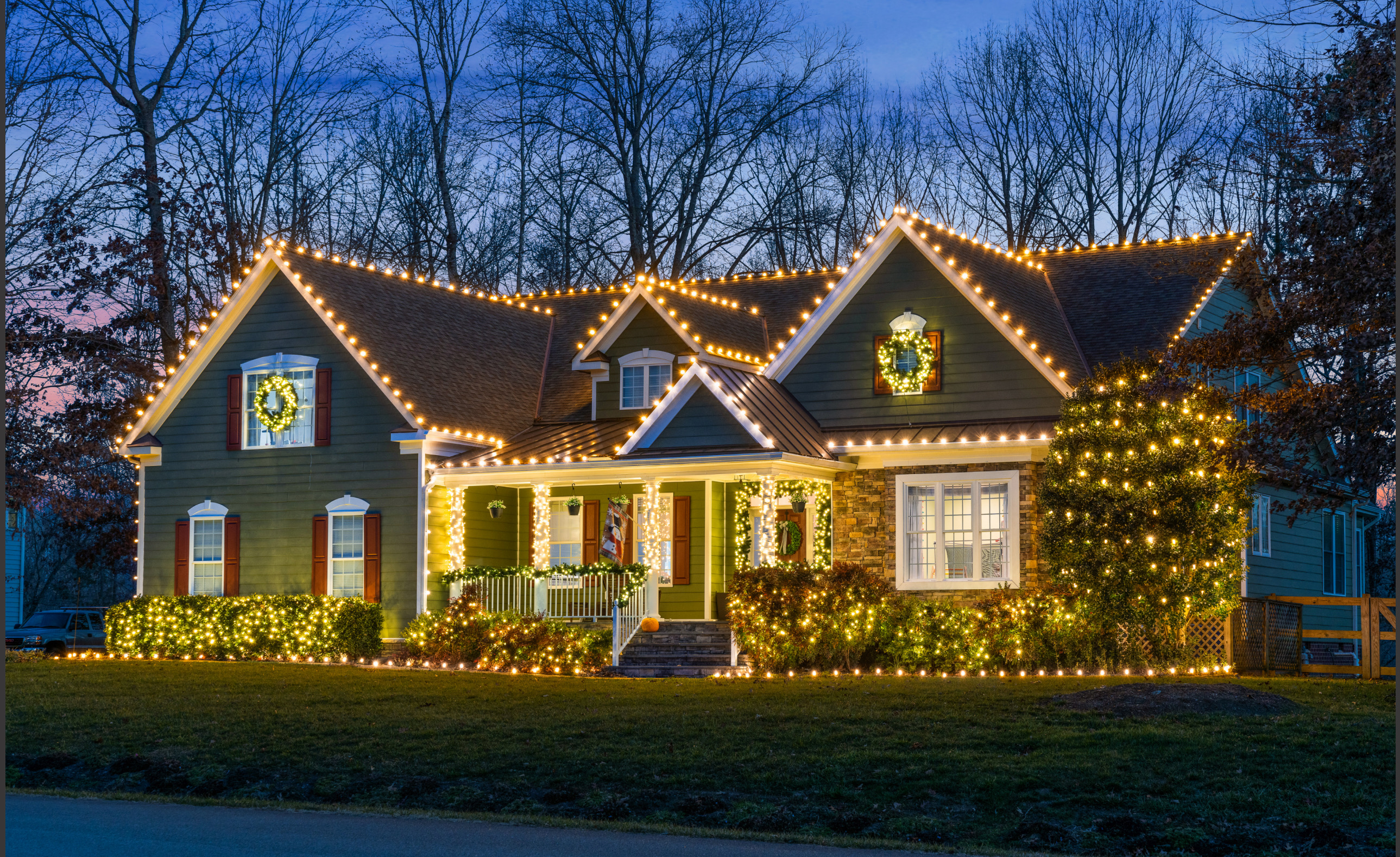 Home with warm white roofline lights, a lit wreath and wrapped trees at dusk