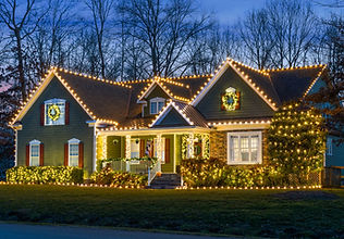 Home with warm white roofline lights, a lit wreath and wrapped trees at dusk