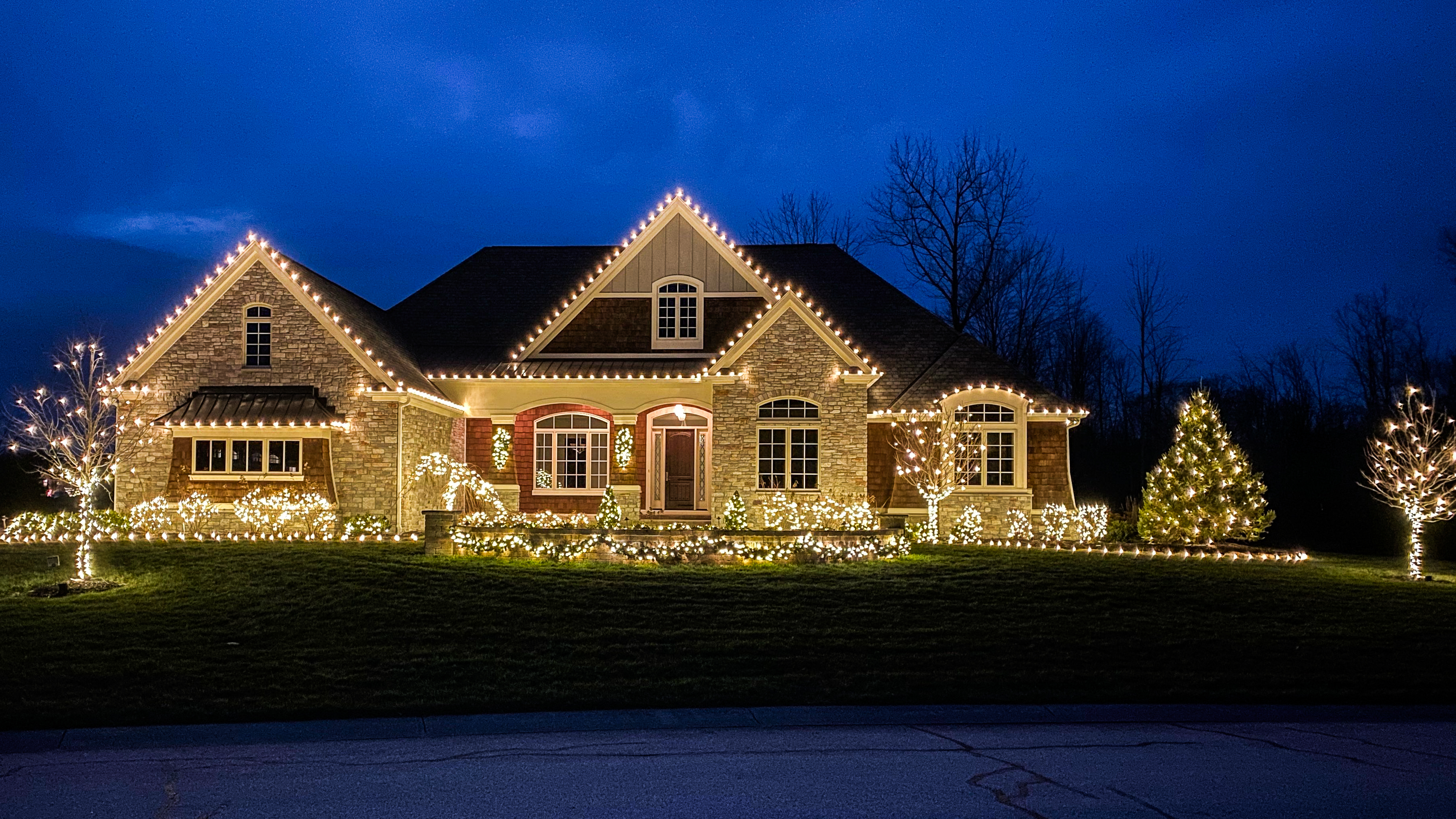 Home roofline, gables and shrubs outlined in warm white Christmas lights at dusk