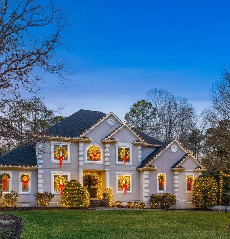 White two-story home with window wreaths and warm white roofline lights at dusk