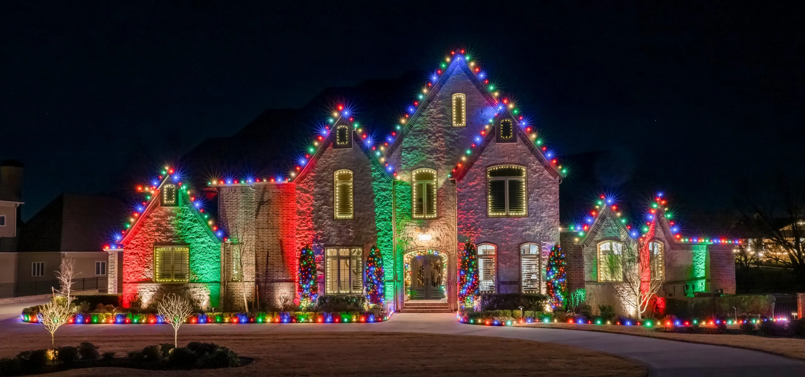 Stone home with red, green and multicolor roofline lights and lit shrubs
