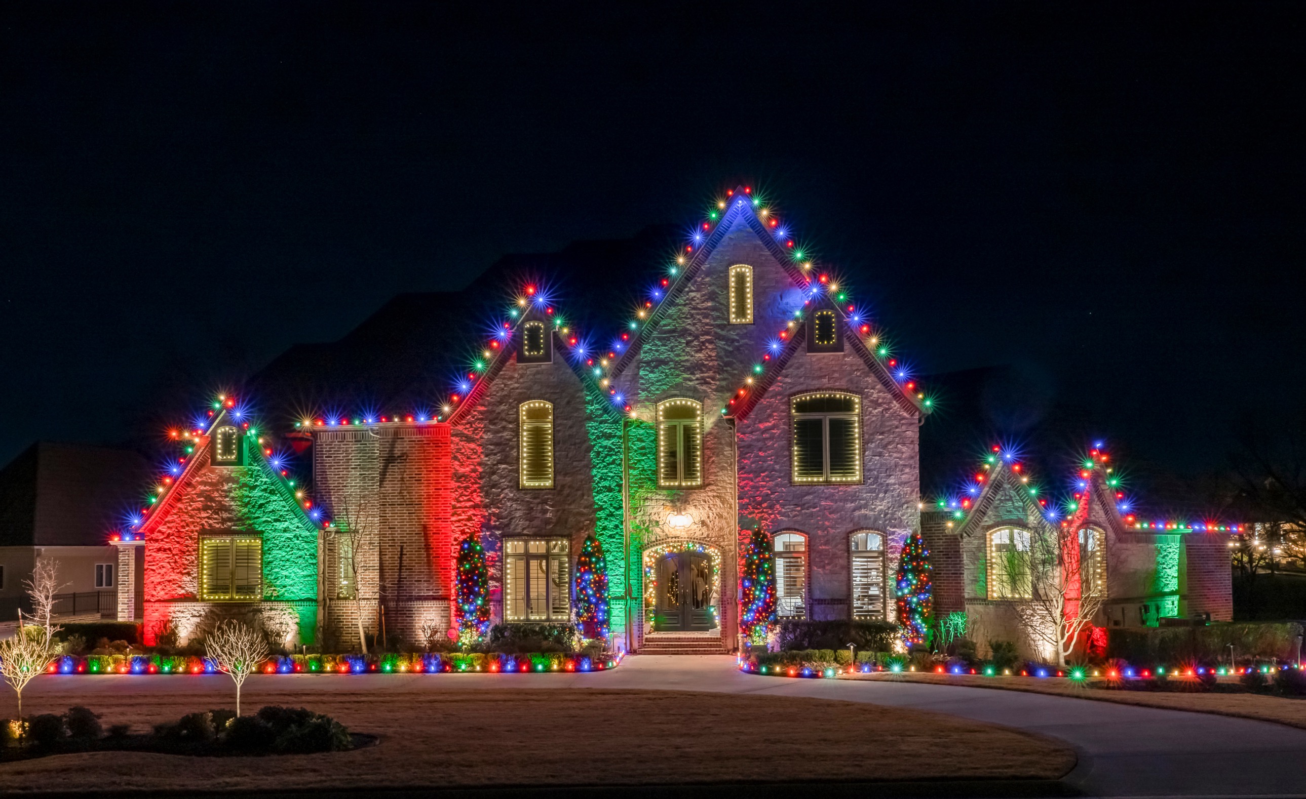Stone home with multicolor roofline lights, colored wall washes and lit shrubs