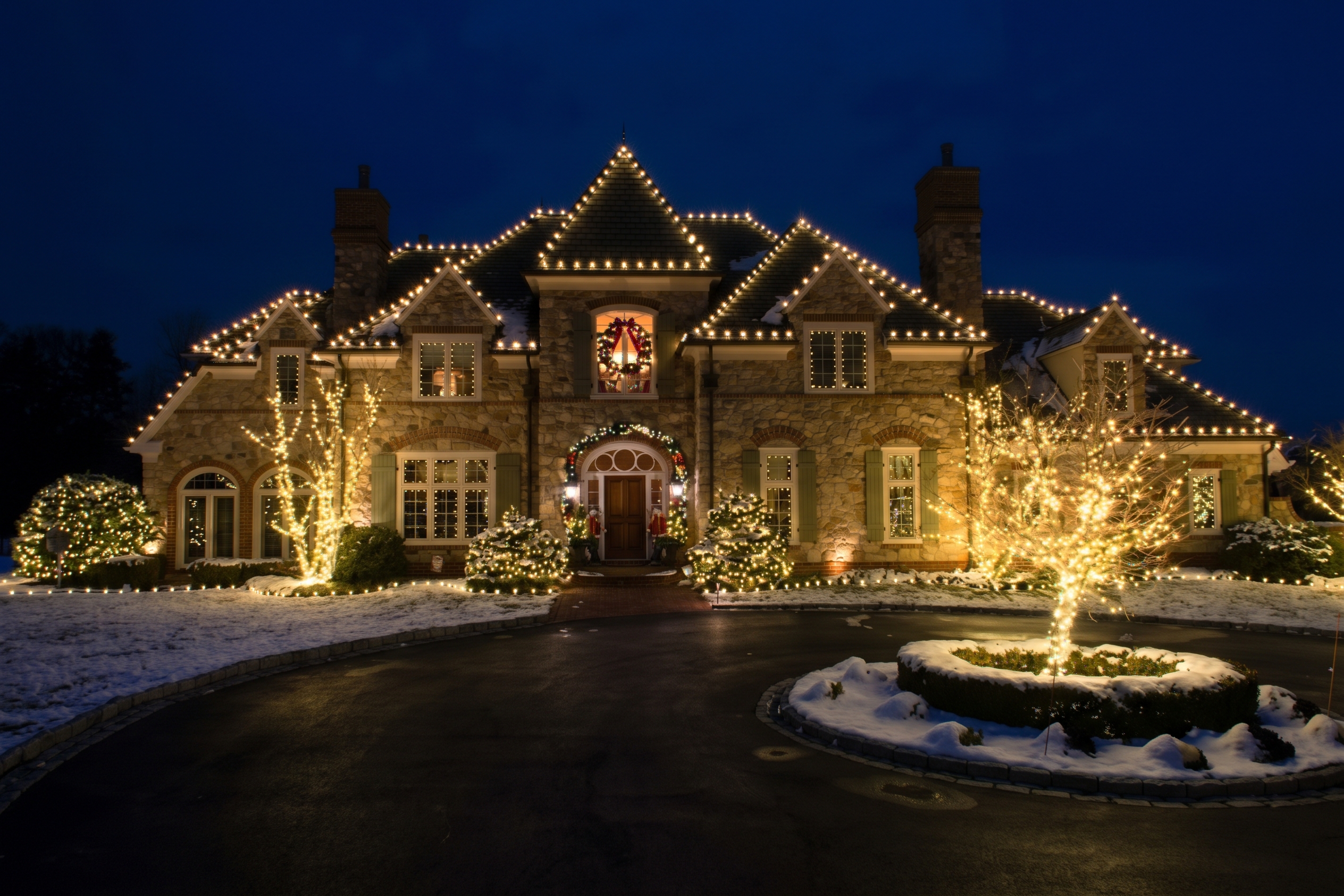 Stone home with warm white roofline lights, lit trees and a lighted fountain