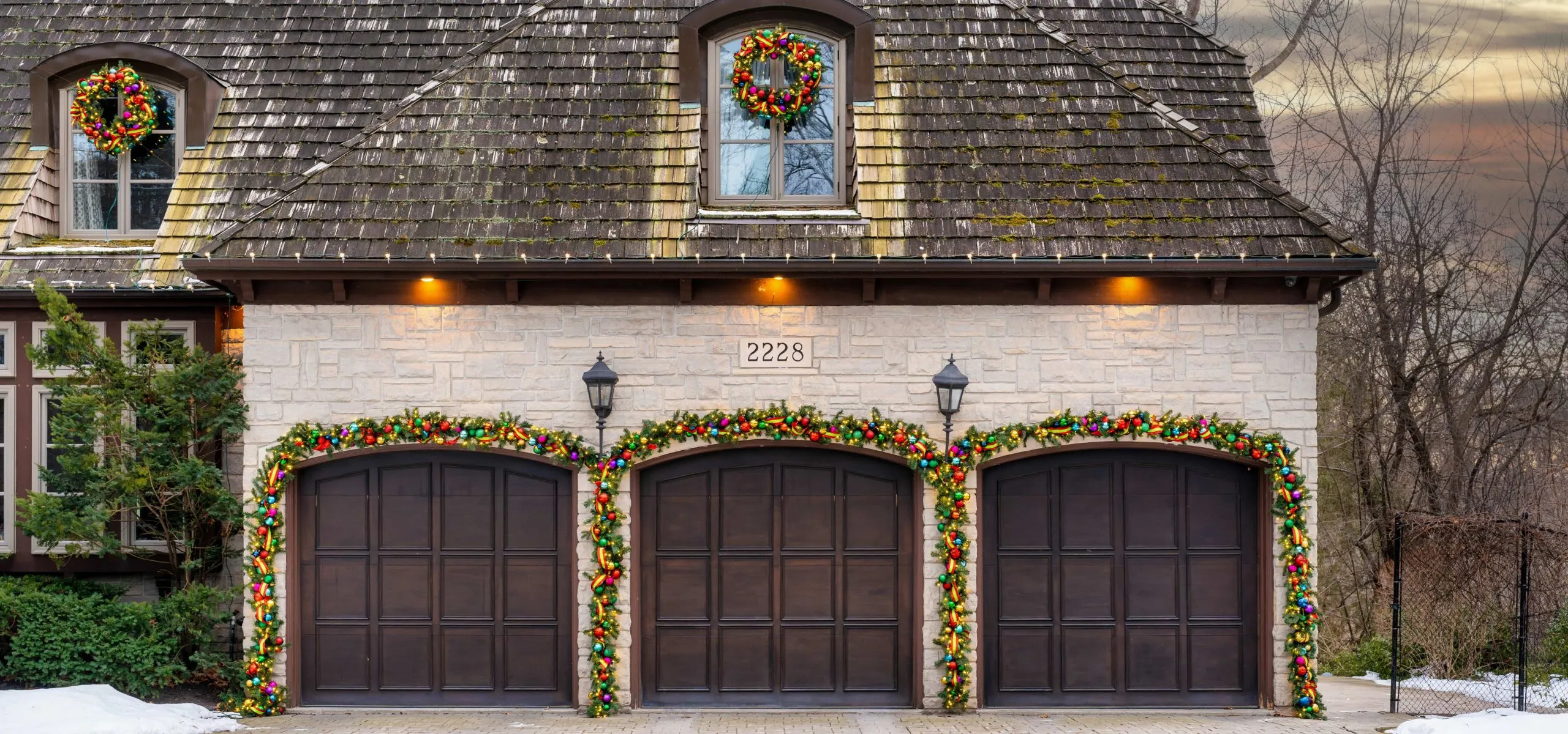 Three-car garage framed with lighted garland and wreaths under roofline lights