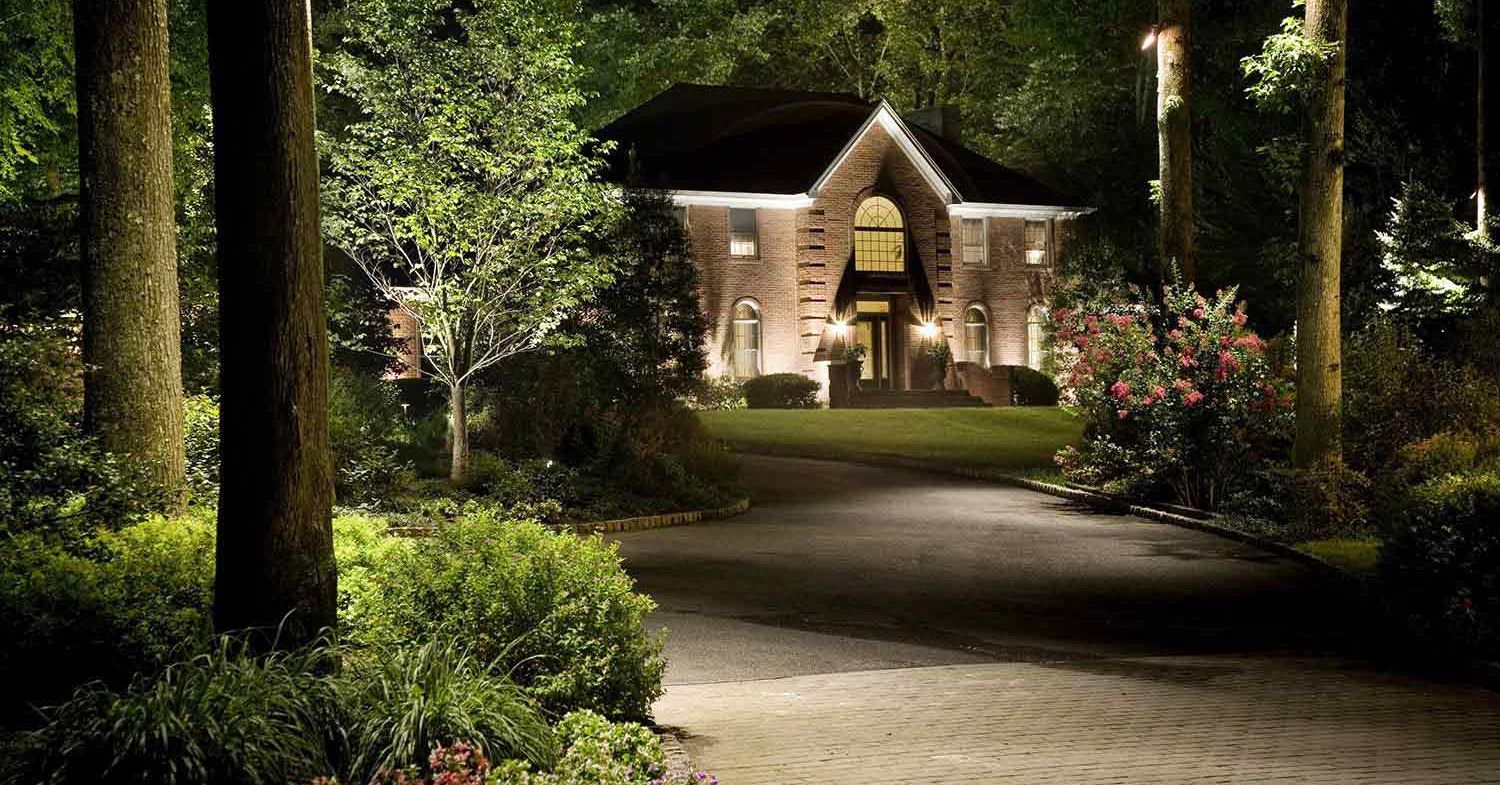 Driveway and lawn with tree uplighting in front of a lit home