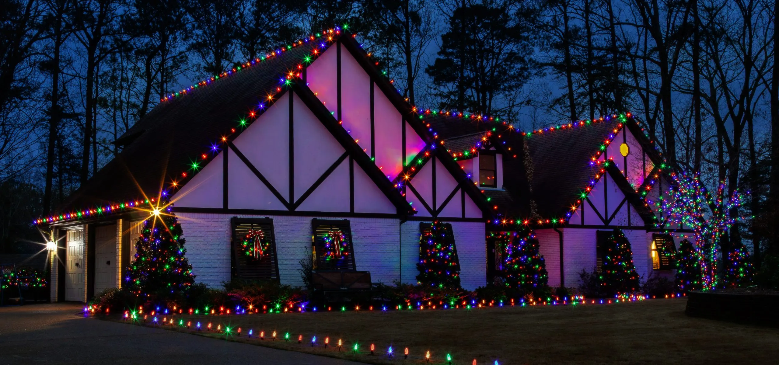 Tudor-style home outlined in multicolor Christmas lights with pink wall washes and lit shrubs