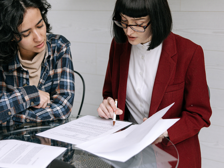 Two women sitting at a table reviewing paperwork together, one pointing to a document while discussing details in a collaborative setting.