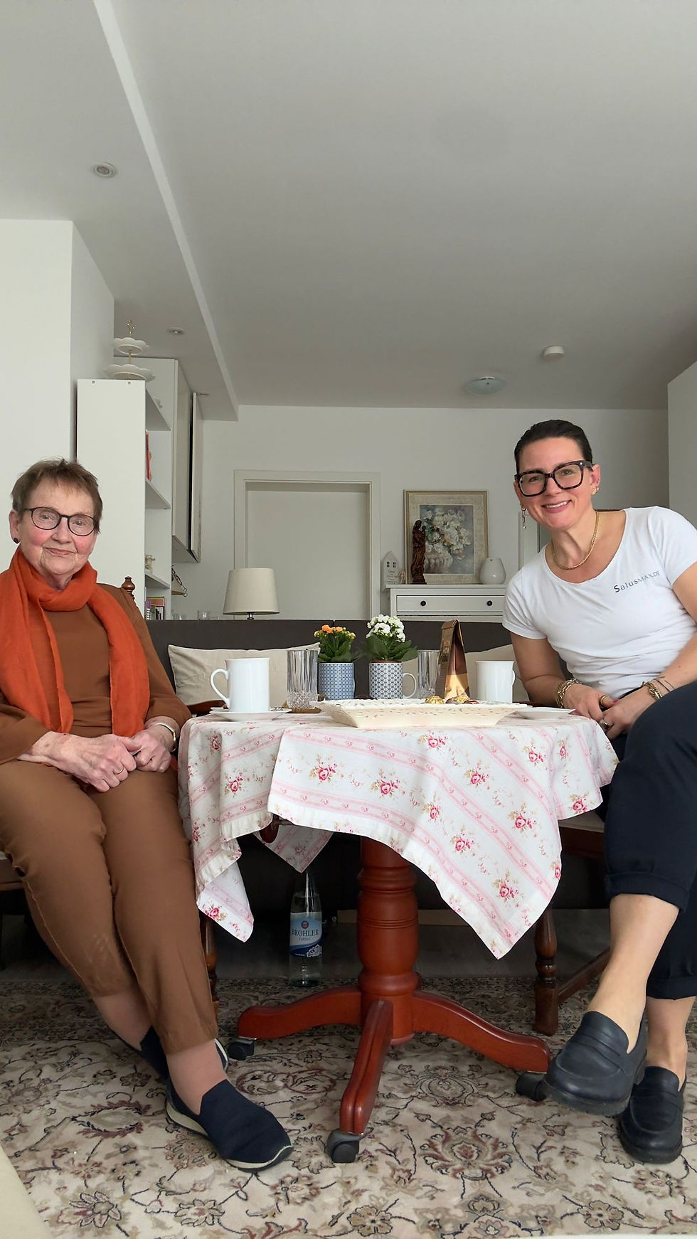 Seniorin sitzt mit SalusMAX Betreuungskraft am gedeckten Kaffee Tisch