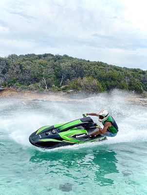Un homme portant un casque et un gilet de sauvetage vert conduit un jet ski Kawasaki vert et noir, effectuant un virage serré et projetant de l'eau sur une mer turquoise près d'une côte rocheuse boisée.