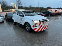 Véhicule d'intervention Isuzu D-Max blanc de Mecano Service FC garé sur l'aire de repos de l'autoroute A36 près de Chemaudin au crépuscule. Le pick-up de dépannage avec ses bandes réfléchissantes rouges et blanches a ses phares allumés, prêt à intervenir sous un ciel gris avec la station service en arrière-plan