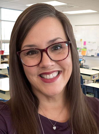 This image of Lisa Combs shows a smiling, middle aged woman with long brown hair and reddish framed glasses standing in the foreground of an empty classroom.