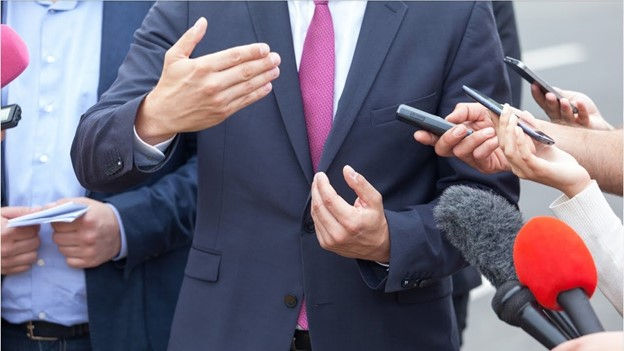 A compelling image featuring a distinguished man dressed in a dark blue suit and a vibrant red tie, engaging in a conversation with a group of reporters. The reporters are holding microphones, indicating their role in conducting interviews and gathering information.