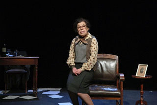 Photographie de scène du spectacle "La vie matérielle", montrant la comédienne Catherine Artigala incarnant Marguerite Duras, assise sur le bras d'un fauteuil en cuir au centre d'un plateau de théâtre sombre comportant un bureau en bois et des feuilles éparpillées au sol.