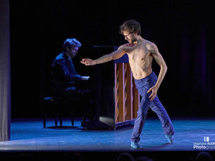 Un danseur torse nu et en pantalon bleu exécute un mouvement expressif au premier plan, la main tendue, pendant qu'un pianiste joue en arrière-plan à côté d'un piano droit ouvert.