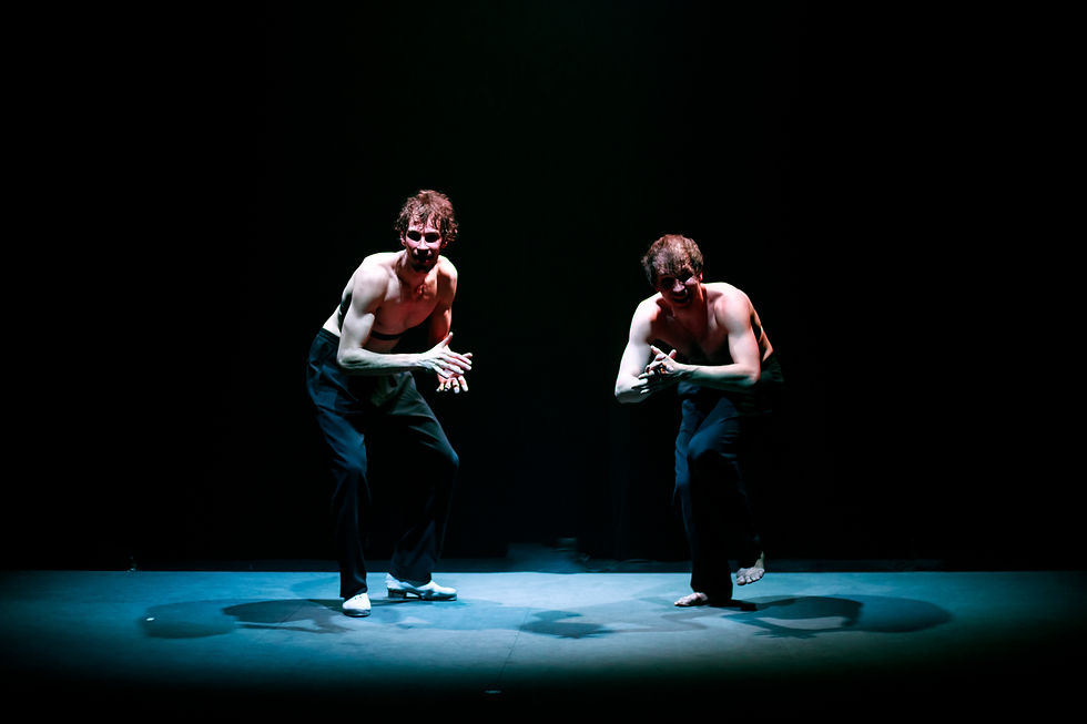 Deux danseurs torse nu et en pantalon sombre adoptent une posture fléchie et expressive au centre d'un halo de lumière bleue sur une scène sombre.