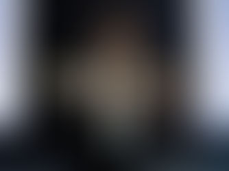 Un comédien en veste noire s'adresse au public la main ouverte au premier plan, devant une scène sombre où une femme est allongée les bras écartés et une autre se tient en arrière-plan.