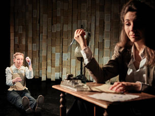 Photographie de scène du spectacle "La Sœur de Shakespeare" par la Compagnie Reme, montrant une comédienne assise au premier plan à un bureau en bois écrivant avec une plume, tandis qu'une seconde comédienne en arrière-plan est assise au sol, le tout devant un mur entièrement tapissé de pages de livres ouvertes.