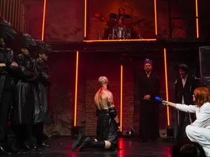 Photographie de scène du spectacle "Hercule" par le Collectif Nox, montrant plusieurs comédiens en costumes sombres et futuristes sur un plateau éclairé par des tubes verticaux orange, avec un batteur installé en hauteur en arrière-plan.
