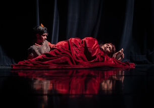 Deux comédiens sur une scène sombre aux reflets mats, l'un veillant au chevet de l'autre allongé et enveloppé dans une imposante couverture rouge.