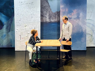Une comédienne assise à une table en bois échange avec un comédien debout à côté d'elle, devant un décor composé de grands panneaux présentant des textes manuscrits et un paysage de montagne.