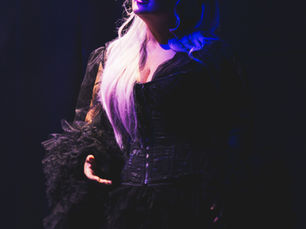 Photographie de scène du spectacle "A queen is born" de Marlène Schaff, mettant en valeur une artiste aux longs cheveux blonds et costume sombre, éclairée par une lumière violette et bleue théâtrale.