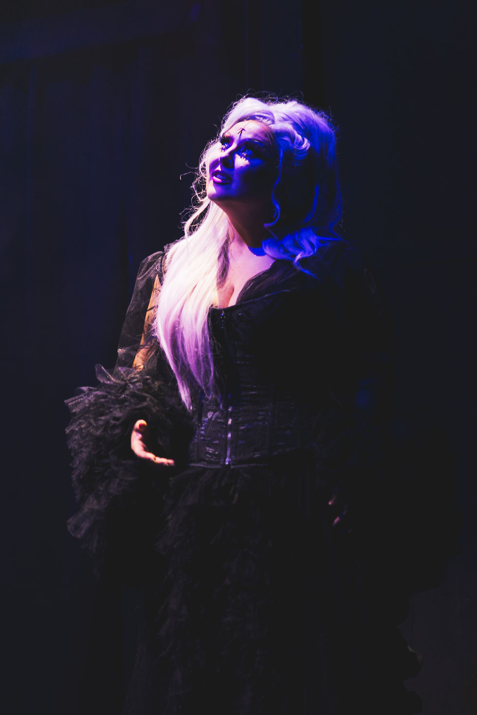 Photographie de scène du spectacle "A queen is born" de Marlène Schaff, mettant en valeur une artiste aux longs cheveux blonds et costume sombre, éclairée par une lumière violette et bleue théâtrale.