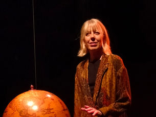 La comédienne Sylvie Dorliat, vêtue d'un long manteau aux motifs orientaux, se tient sur une scène sombre à côté d'un grand globe terrestre suspendu.