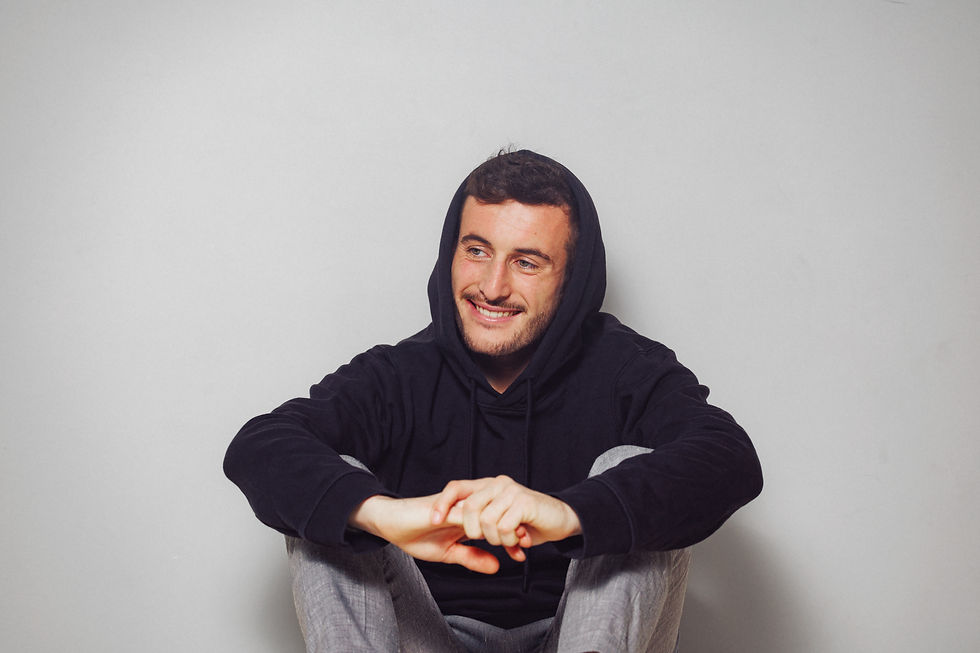 Portrait en plan moyen du comédien Samuel Certenais assis sur fond gris clair uni, portant un sweat à capuche noir relevée sur la tête et un pantalon gris, affichant un sourire détendu en regardant vers le côté.