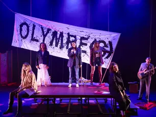 Cinq comédiennes posent sur et autour d'une praticable de scène devant une grande banderole blanche peinte du titre « OLYMPE(S) », accompagnées d'un musicien jouant de la guitare électrique sur le côté.