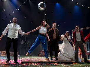 Photographie de scène du spectacle "Le jeu de l'amour et du hasard", montrant six comédiens tout sourire se tenant par la main en ligne sur un plateau coloré de confettis, sous des guirlandes lumineuses et une boule à facettes, avec des bottes de paille en arrière-plan.