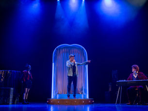 Photographie de scène du spectacle "Felicita", montrant un comédien au centre debout sous une arche lumineuse bleue, entouré à gauche d'une comédienne près de loges de théâtre et à droite d'un musicien assis derrière son clavier, le tout baigné dans une forte lumière scénique bleue.
