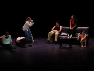 Photographie de scène du spectacle "Écoutez leur silence" de Gabrielle Gay, montrant six comédiens répartis sur un plateau sombre, certains assis sur un canapé noir au centre, d'autres au sol ou debout dans des postures d'attente et d'incompréhension.