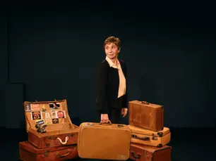 La comédienne Ariane Ascaride, vêtu d'une veste sombre et d'une chemise claire, se tient au centre d'un décor composé de plusieurs valises anciennes empilées et ouvertes.