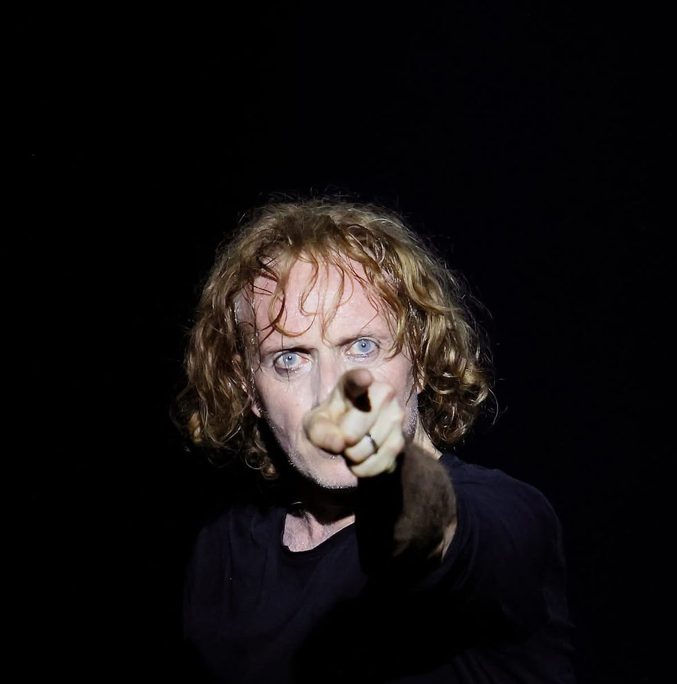 Photographie de scène en clair-obscur montrant le comédien William Mesguich sur fond noir, le visage éclairé par un faisceau de lumière, fixant intensément l'objectif et pointant fermement le doigt vers l'avant.