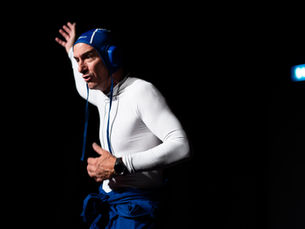 Photographie de scène en clair-obscur montrant le comédien Yohann Métay en haut blanc moulant et bonnet de bain bleu, le bras levé et le regard expressif, se détachant sur un fond noir de théâtre.