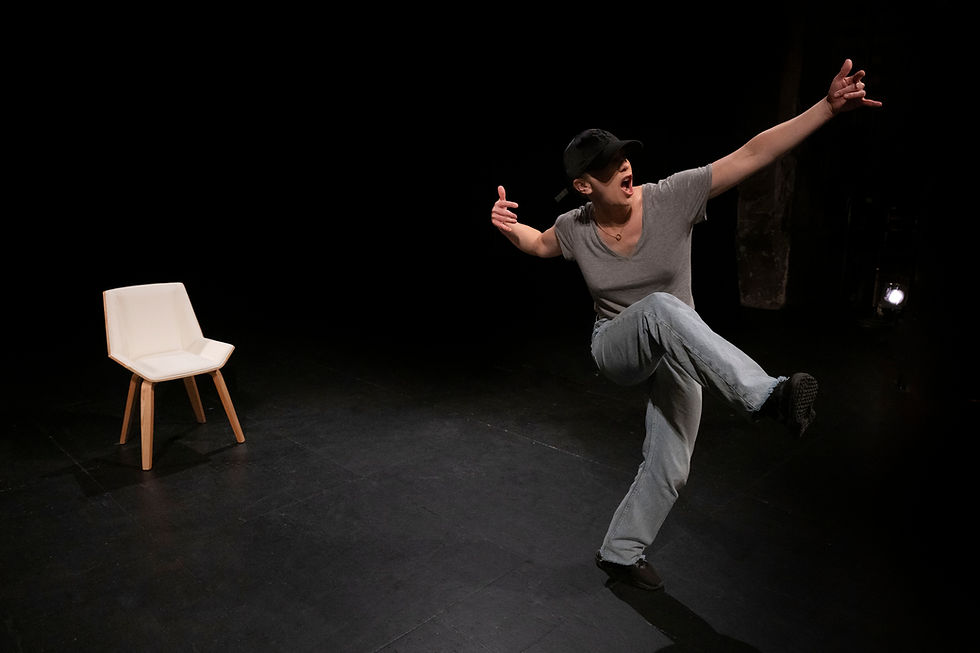 Une comédienne casquée, en tee-shirt gris et jean, effectue une posture de danse dynamique et expressive sur une scène sombre, à côté d'une chaise blanche.