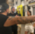 People socializing on a street at night; a tattooed arm extends towards a smiling man and a woman in white shorts. Bright yellow storefront.