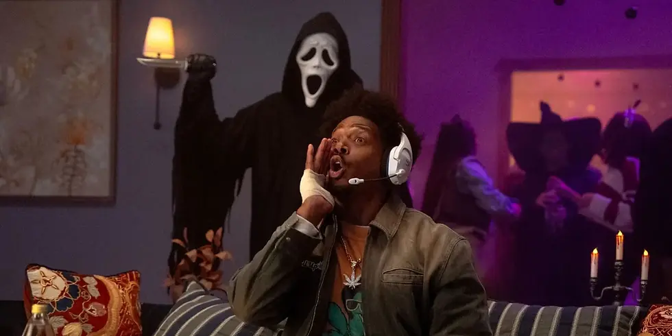 Szenenbild aus „Scary Movie“: Ein Mann mit Gaming-Headset ruft erschrocken, während sich hinter ihm eine schwarz gekleidete Gestalt mit weißer Maske und Messer nähert.